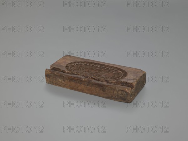 Double-sided gingerbread mould. Obverse: Madonna and Child—Madonna wearing a crown, in a long patterned dress, surrounded by a double oval inner frame forming tongues of flame and an outer laurel-wreath frame. Reverse: two standing figures in richly decorated 17th–18th-century court costumes a man with a long wig, justaucorps, culottes and stockings; a woman with a wig, long dress with a neckline, skirt decorated at the bottom with a plant tendril. Materials/technique: gingerbread mould; decorative applied art with plant ornaments, religious motifs and symbols. Date: 18th–19th century. Place/style: Silesia Province of Silesia, 1742–1918. Dimensions: height 20.3 cm, width 10 cm, depth 3.5 cm., A rectangular wooden block with a carved oval recess sits on a smooth grey surface; the block shows worn and cracked edges, rough texture, and visible wood grain. The carving inside the oval has detailed, raised patterns and a scalloped border. The wood displays various shades of brown, including ...