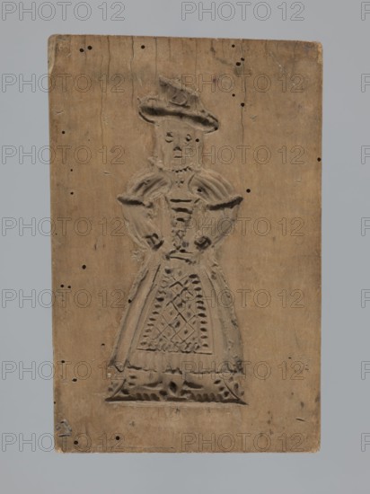 Double-sided gingerbread mold; obverse: female figure; reverse: four images baby in swaddling clothes, fish, two shoes. Gingerbread, applied/folk decorative art; figurative and zoomorphic motifs; female figures in rich patterned dress and hat. Silesia Province of Silesia, 18th–19th century, 1742–1918. Rectangular form; height 17 cm, width 11 cm, depth 2 cm., A rectangular wooden board with a carved shallow relief of a standing figure wearing a hat and a long dress, the carving showing details of a bodice, puffed sleeves, a patterned apron, and hands resting at the waist; the wood surface is light to medium brown with darker brown grain lines, small holes, and faint scratches; the background behind the board is a flat pale gray.