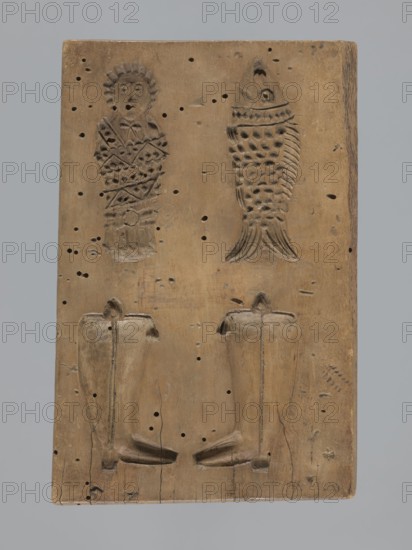 Double-sided gingerbread form. Obverse: female figure in a long patterned dress and a hat. Reverse: four images — a baby in swaddling clothes, a fish, and two shoes. Gingerbread mold, applied/folk decorative art with figurative and zoomorphic motifs; female figures in rich dresses; folk motifs. Rectangular form. 18th–19th century. Province of Silesia 1742–1918. Height 17 cm, width 11 cm, depth 2 cm., A rectangular wooden board with a warm tan-brown surface showing natural grain and darker brown areas, small dark holes and scattered small black pinpricks, and some lighter worn spots; the board bears carved recessed shapes including a human-like figure at the upper left with a rounded head, carved facial features, and a textured patterned body, a fish-shaped carving at the upper right with carved scales and a tail, and two boot-shaped or vessel-like vertical molds at the lower half with raised tops and curved bases; the edges of the board are slightly darker and the bottom edge shows a t...
