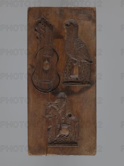 Double-sided rectangular gingerbread mold with three representations on each side. One side: rooster, ox, and dog arranged vertically on bases, decorated with geometric and floral motifs. Other side: guitar, stork, and rider on a horse blowing a horn and wearing a bicorn, each motif embellished with geometric-floral ornament. Zoomorphic and figural motifs; folk decorative art. Material: gingerbread mold form. Date: 18th–19th century. Region: Province of Silesia 1742–1918. Dimensions: height 16.7 cm, width 7.7 cm, depth 3.2 cm., A rectangular wooden panel with a brown surface featuring carved reliefs, including a stringed instrument shape, a bird perched on a decorated pedestal, and a rider on a horse on a decorated base; the wood shows darker and lighter brown variations and some glossy wear, with the background around the panel a muted gray.