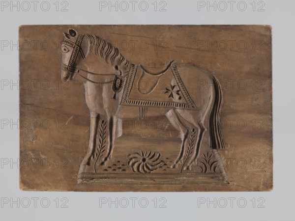 Double-sided gingerbread mould. Rectangular shape; one side: stylised horse in full harness with a Maltese cross on the saddle, standing on a base decorated with floral and plant ornament; other side: deer eating leaves, with an ornament of leaves and flowers at the bottom. Folk-style decorative motifs; applied decorative art for gingerbread. 18th–19th century, Province of Silesia 1742–1918. Height 15.5 cm, width 10.2 cm, depth 3.4 cm., A carved wooden panel depicting a horse in profile standing on a rectangular base, with detailed carved mane, tail, bridle, and a decorative saddle cloth; the carving shows stylized floral and leaf motifs beneath and around the horse and textured decorative borders on the saddle cloth and bridle. The panel and carving are primarily brown with variations in wood tone, and the background area around the panel appears light gray.