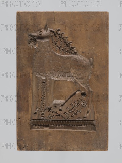 Double-sided gingerbread mould. Rectangular shape; obverse: stylised horse in full trot with a Maltese cross on the saddle, standing on a base decorated with floral and plant ornament; reverse: deer eating leaves, with a leaf-and-flower ornament at the bottom. Folk-style decorative motifs; applied/decorative art object used for gingerbread. 18th–19th century, Province of Silesia 1742–1918. Materials: gingerbread mould; dimensions: height 15.5 cm, width 10.2 cm, depth 3.4 cm., A carved wooden plaque showing a stylized animal in profile with a long neck and body, standing on a carved base decorated with floral and leafy motifs; the animal has a textured coat indicated by repetitive carved lines, a detailed mane made of braided or looped carved elements, a small rounded tail, and a leafy branch or foliage held in its mouth. The plaque surface is worn with visible tool marks and small holes, and the colors visible are various shades of brown on the wooden plaque set against a pale gray bac...