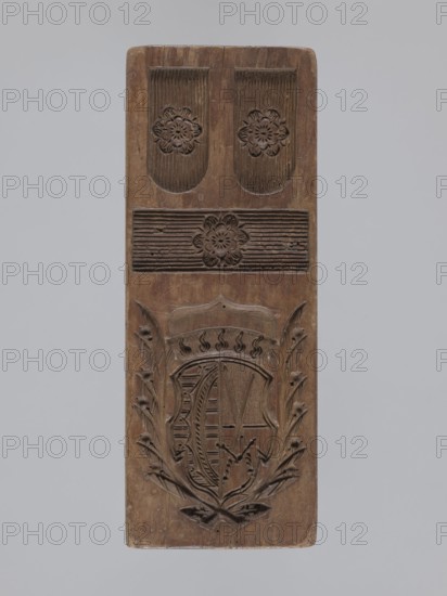 Double-sided gingerbread form. Obverse: heraldic shield; reverse: plant ornament. Double-sided rectangular form; on one side at the top a heraldic shield with a bipartite field: left, two crossed swords; right, geometric and plant motifs including an image of rue. In the lower part three framed fields, one rectangular, the other two ending with an arc, each containing on a dashed background a rose symbol. Reverse: complex decorative motif of symmetrically arranged and interwoven plant elements. Materials/technique: form. Iconography: coats of arms, roses, plant ornaments, geometric ornaments, swords, crown motif, rue, grapevine. Function: gingerbread mold. Region/date: Silesia, Province of Silesia 1742–1918; 18th–19th century. Dimensions: height 26.7 cm; width 10.2 cm; depth 3.8 cm., A tall rectangular wooden panel with carved decorative motifs, showing a brown wood tone with lighter and darker variations; the top section has two rounded-rectangular recessed panels each containing a ca...