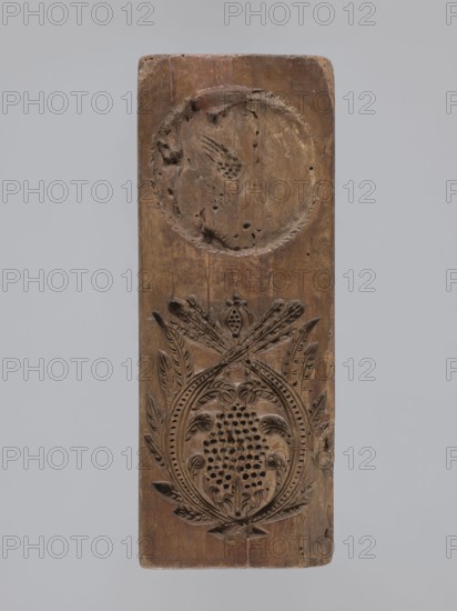 Double-sided gingerbread form. Obverse: heraldic shield; reverse: plant ornament. Double-sided rectangular form; obverse top has a heraldic shield with a bipartite field: left side two crossed swords; right side geometric and plant motifs including rue; lower part three framed fields—one rectangular, two arched—each containing a rose on a dashed background. Reverse: complex decorative motif of symmetrically arranged, interwoven plant elements. Materials: form applied art. Motifs: coats of arms, roses, plant and geometric ornaments, swords, crown, grapevine. Date: 18th–19th century. Place/region: Province of Silesia 1742–1918. Dimensions: height 26.7 cm; width 10.2 cm; depth 3.8 cm., A vertical wooden panel with a warm brown tone sits against a pale gray background, showing a carved circular recess near the top with an irregular carved motif inside and a larger ornate oval carving below featuring a patterned central area surrounded by stylized leaves and curved decorative elements, with...