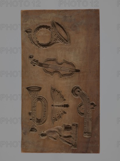 Double-sided gingerbread form. Obverse: four representations — two richly decorated oriental pipes and two long shoes decorated with a floral ornament. Reverse: six different motifs — a human figure sitting on a bucket, a butterfly, a trumpet, a pistol, a violin and a French horn. Materials/technique: applied art; folk motifs. Style/region/date: Silesia Province 1742–1918, 18th–19th century. Form: double-sided rectangular form. Dimensions: height 25 cm, width 11.2 cm, depth 2.2 cm. Creator: unknown., A rectangular wooden board in brown with carved recesses showing a few decorative shapes including a curved horn-like shape near the top, an elongated stringed instrument shape in the upper middle, an oval ornate shape with a tubular element in the lower left area, a winged insect-like form near the center, a stylized human figure reclining on a small platform toward the bottom, and a few scroll-like ornamental elements on the right; the background is plain and the visible colors are shade...