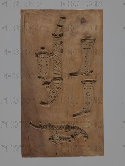Double-sided gingerbread form. Obverse: four representations — two richly decorated oriental pipes and two long shoes decorated with a floral ornament. Reverse: six different motifs — a human figure sitting on a bucket, a butterfly, a trumpet, a pistol, a violin and a French horn. Materials: applied art; form: double-sided rectangular. Style/region: folk motifs, Silesia Province, 18th–19th century. Dimensions: height 25 cm, width 11.2 cm, depth 2.2 cm., A rectangular wooden panel with carved impressions showing decorative boot shapes and ornamental curved tools, the wood surface in shades of light brown and tan with darker brown carved recesses and a few small dark brown knots and cracks, set against a neutral gray background.