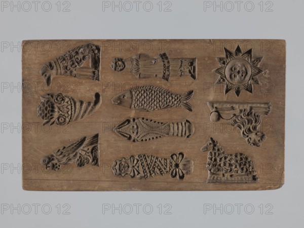 One-sided gingerbread form with various representations, woodcarver or gingerbread maker, gingerbread form, figurative motifs, folk-style decorations, cornucopia ornament, dates, applied art, holiday traditions, lamb symbol, zoomorphic motifs, Silesia Province of Silesia, 1742–1918, babies, souvenirs. One-sided rectangular form with ten small images: bird, cornucopia, lamb, baby in swaddling clothes, crayfish, fish, girl in a dress, another lamb, lion, sun; on one edge a date engraved 1810 with the letter S against an anchor maker’s mark. 18th–19th century. Form: height 20.6 cm, width 12 cm, depth 4 cm., A rectangular wooden board with carved compartments containing a group of decorative relief shapes, shown against a neutral gray background; the board is brown with visible wood grain and wear, and contains a few carved figures including a sunburst with a face, a fish with textured scales, a lamb with a patterned fleece, a horn-shaped design with floral motifs, a cornucopia-like carved...