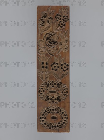 Double-sided gingerbread mould. Oblong rectangular shape. Obverse: 7 floral and plant motifs — bouquets of roses and other flowers and two floral wreaths. Reverse: 12 various motifs in frames and medallions — a sequence of twelve images in a rectangular frame depicting a human figure during farm work, below eight medallions containing representations of human heads and various animals. Materials: gingerbread mould for gingerbread, applied art; folk-style decorations, figural scenes, floral and zoomorphic motifs. Date: 18th–19th century. Place: Province of Silesia 1742–1918. Dimensions: height 25.3 cm, width 6.6 cm, depth 1.5 cm., A vertical rectangular wooden panel with carved floral designs running along its length, showing a carved rose and leaves near the top, carved small flowers and leaves arranged in wreath-like circular patterns toward the middle and bottom, and carved leaf motifs interspersed among the flowers; the wood appears brown with variations in tone and the background i...
