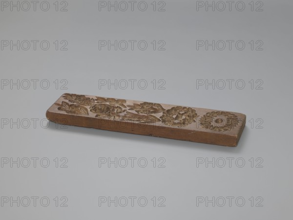 Double-sided gingerbread mould. Obverse: 7 floral and plant motifs; reverse: 12 various motifs in frames and medallions. Rectangular mould with one side showing bouquets of roses and other flowers and two floral wreaths; second side a sequence of twelve images in a rectangular frame depicting a human figure during farm work, below eight medallions containing representations of human heads and various animals. Gingerbread mould with folk-style decorations, wreaths, human figures, floral and zoomorphic motifs; applied art, figural scenes. 18th–19th century, Province of Silesia 1742–1918. Height 25.3 cm; width 6.6 cm; depth 1.5 cm., A rectangular wooden block rests on a smooth surface, showing carved floral and circular wreath-like patterns on its top face; the wood displays warm brown tones with some darker carved recesses and slightly lighter worn edges, and the background and surface are shades of pale gray.