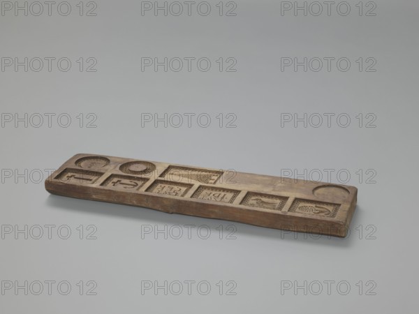 Double-sided gingerbread mould. Obverse: images of four heads; depending on rotation the heads take on a male or female appearance in one the male head changes into a goat. Reverse: seven elements in decorative frames: a house roof, a flower, a bird, a plate with the inscription IDR / 1763 and another with GFK / 1763, an anchor and cross, decoration complemented by a wreath and flower motif in a medallion. Plant ornaments, human figures, zoomorphic motifs, wreaths, religious symbols. Applied/folk art, decorative art. Date: 1793. Maker/ Region: Province of Silesia 1742–1918. Form. Dimensions: height 34.6 cm, width 9.3 cm, depth 2 cm., A long rectangular wooden board rests on a smooth light gray surface, with a muted gray background. The board is brown with variations of lighter and darker wood tones and shows carved recessed panels and circular indentations along its top face. Several square and rectangular carved compartments display raised decorative reliefs and patterned borders, and...