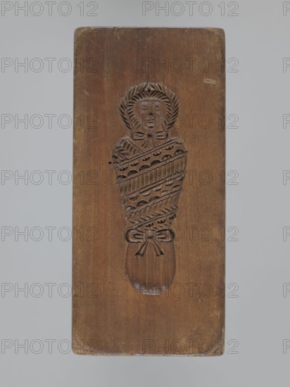 Double-sided gingerbread mold. Obverse: baby in swaddling; Reverse: baby in swaddling. Folk-style decorations with geometric and floral ornament; decorative frame around the figure. Applied art, decorative art, souvenir gingerbread mold. 18th–19th century. Province of Silesia 1742–1918. Mold; height 16.8 cm, width 7.8 cm, depth 3.7 cm., A rectangular wooden board in shades of brown with a carved relief of a wrapped figure centered on it; the carving shows a face encircled by a textured halo, a bow beneath the chin, patterned wrapped layers around the body, and a bow near the lower end, with the wood surface showing grain, slight scratches, and varied brown tones against a gray background.