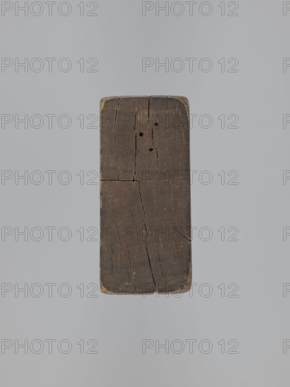 Double-sided gingerbread mold; obverse: baby in swaddling; reverse: baby in swaddling. Rectangular mold with richly decorated geometric and floral ornament and a decorative frame around the figure. Folk-style decorative art, souvenirs, gingerbread mold. 18th–19th century, Province of Silesia 1742–1918. Mold; height 16.8 cm, width 7.8 cm, depth 3.7 cm., A rectangular block of aged wood with a dark brown surface suspended against a light gray background, showing a few cracks and splits running along its grain, a few small round holes near the top, worn rounded corners with lighter tan edges, and subtle traces of reddish discoloration on the face.