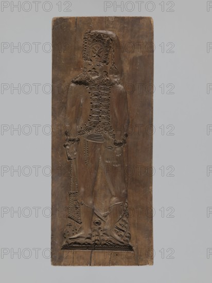 Double-sided gingerbread form. Obverse: male figure with a moustache in a hussar uniform, wearing a fur-lined jacket, standing with legs slightly apart, leaning on a sabre held in his right hand, wearing a high shako topped by a two-headed eagle and tassel; plant ornament at the bottom. Reverse: stylized elongated fish covered with scales, depicted in profile. Rectangular form, applied art/gingerbread mold, decorative folk motifs, soldiers/hussars, historical costumes. 18th–19th century, Province of Silesia 1742–1918. Height 28.7 cm; width 11.7 cm; depth 3.5 cm., A carved wooden panel in brown tones shows a relief of a standing human figure with detailed textured hair and a tall hat, wearing a textured upper garment and a belt, with visible sculpted hands and feet, and holding a long object that reaches to the base; the panel background is smooth with variations in brown, and the image also shows a pale gray background surrounding the panel.