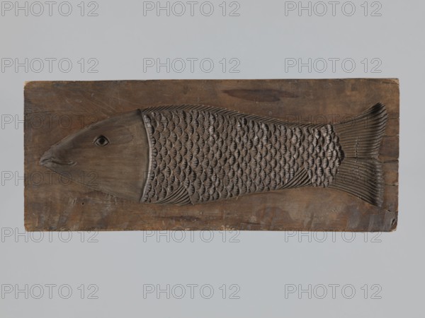 Double-sided gingerbread form. Obverse: male figure in a hussar uniform; reverse: fish. Double-sided rectangular form; on one side an image of a male figure with a moustache in a hussar uniform, wearing a fur-lined jacket, standing with legs slightly apart, leaning on a sabre held in his right hand, wearing a high shako topped with a two-headed eagle decorated with a tassel; plant ornament at the bottom. Other side: stylized elongated fish covered with scales, depicted in profile. Materials/function: gingerbread form, applied/ decorative art; motifs: soldiers, hussars, uniforms, sabres, folk motifs, fish ornament, historical costumes. Date: 18th–19th century. Place: Province of Silesia 1742–1918. Dimensions: height 28.7 cm; width 11.7 cm; depth 3.5 cm., A carved wooden relief of a fish mounted on a rectangular wooden panel, showing a stylized fish with a smooth head and a textured body of repeating scale patterns, a carved eye and mouth, and a detailed tail with linear ridges; the pane...