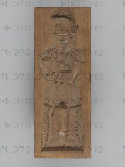 Double-sided gingerbread form; obverse: female figure in a helmet; reverse: male figure in a helmet. Gingerbread mold, applied/decorative art, militaria, historical costumes, plant ornaments. Rectangular form; on one side a female figure in a long patterned dress with a neckline, wearing a helmet decorated with a plume, holding a sword in one hand and a roll of paper in the other, wearing shoes, with a plant ornament at the bottom. The other side shows a male figure in armor resembling Roman legionary or Greek hoplite armor, wearing a similar plume-decorated helmet, holding a shield bearing two crossed swords and a crown in one hand and a javelin in the other, a sword girded at his side, wearing sandals, with a plant ornament at the bottom. 18th–19th century, Silesia Province 1742–1918. Height 59 cm, width 22 cm, depth 4.5 cm., A carved wooden panel depicting a standing armored figure wearing a helmet with a crest and a plume, holding a spear in one hand and a round shield in the other...