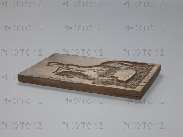 Double-sided gingerbread form. Obverse: image of a rider. Reverse: four representations of human figures. Double-sided rectangular form; on one side an image of a trumpeter on a horse dressed in a decorative tail, in a costume from the turn of the 18th and 19th centuries consisting of a long jerkin with wide tails, breeches and long boots, wearing a tricorn hat; floral decoration at the bottom. Second side: four small images of human figures shown from the front and back: first, a male carrying a basket filled with children probably known in folk tradition as Kinderbringer; second, a female in a high headdress, in country dress, holding a handkerchief in one hand and a small basket in the other. Gingerbread form, 18th–19th century, Province of Silesia 1742–1918. Dimensions: height 42 cm, width 25.5 cm, depth 2.8 cm., A flat rectangular wooden block rests on a smooth light gray surface against a pale gray background, showing carved raised and recessed patterns across its top face; the w...
