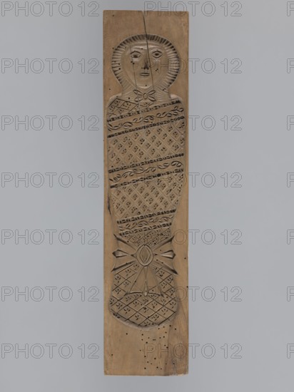 Double-sided gingerbread mold. Obverse: baby in swaddling clothes richly decorated with various geometric and plant motifs. Reverse: two rectangular frames filled with geometric ornaments. Double-sided rectangular mold, gingerbread; applied/decorative arts, folk motifs, folk customs; human figures, plant and geometric ornaments. 18th–19th century, Province of Silesia 1742–1918. Height 40.5 cm; width 9.5 cm; depth 2.3 cm., A vertical wooden plank carved in a light brown wood tone features a tall carved figure with a round head and simple facial features, a textured halo or hood around the head, a wrapped body decorated with diagonal patterned bands and repeated small diamond motifs, and a tied bow or knot shape near the lower section; the carving shows tool marks and small holes, and the background around the plank is a soft grey.