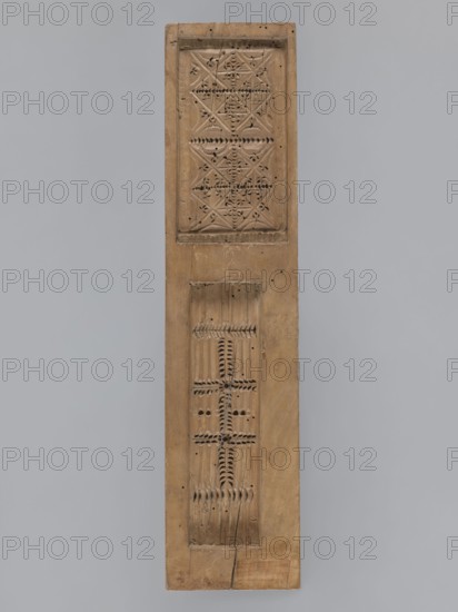 Double-sided gingerbread mold. Obverse: baby in swaddling clothes richly decorated with geometric and plant motifs. Reverse: two rectangular frames filled with geometric ornaments. 18th–19th century. Province of Silesia 1742–1918. Mold; height 40.5 cm, width 9.5 cm, depth 2.3 cm. Materials/technique: applied arts; decorative folk motifs include geometric and plant ornaments, human figure; function: gingerbread mold., A long vertical wooden panel with a natural tan and brown wood grain, showing a few carved rectangular recessed areas with decorative carved patterns; the top recessed area contains a geometric arrangement of repeating carved shapes and small pierced holes, while the lower recessed area shows a series of linear carved grooves and repeated notched motifs with several small drilled holes; the panel has visible wood texture, some small dark spots and cracks, and overall warm beige and brown tones set against a smooth light gray background.