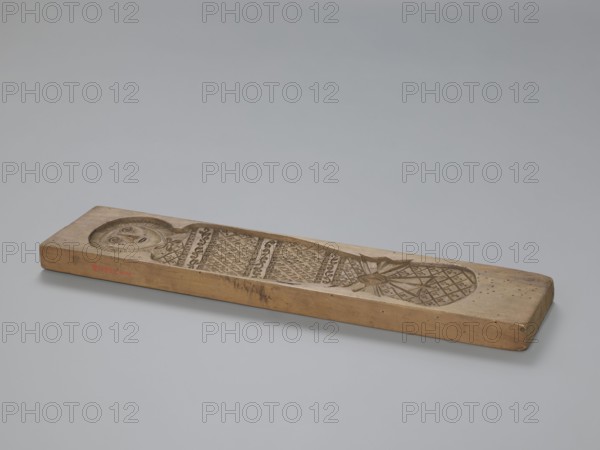 Double-sided gingerbread mold. Obverse: baby in swaddling clothes richly decorated with various geometric and plant motifs. Reverse: two rectangular frames filled with geometric ornaments. 18th–19th century. Province of Silesia 1742–1918. Mold; height 40.5 cm, width 9.5 cm, depth 2.3 cm. Materials: gingerbread mold. Subjects: babies, geometric ornaments, plant ornaments, human figures, folk motifs, folk customs., A long rectangular wooden plank rests on a smooth light gray surface, carved along its top with an elongated recessed design depicting a stylized human face at one end and a patterned, lattice-like section toward the other end, connected by bands of repeating ornamentation; the wood shows natural tan and brown tones with areas of darker brown where the carvings and edges have aged, and there are a few small darker spots and scuffs on the plank.