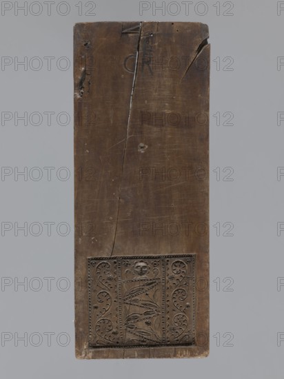 Double-sided gingerbread form. Obverse: figure of an angel with the alphabet. Reverse: various floral ornaments. Double-sided rectangular form; on one side a male figure probably an angel standing, in a costume richly decorated with plant ornament, girded with two sashes, holding a decorative board with the alphabet inscribed; above the figure's head a halo; in the background a feather and flame motif possible angel wings. The other side: rectangular field filled with floral and plant decoration, including the head of a baby. At the top a signature: initials G and R mark of the woodcarver or gingerbread maker/workshop owner. 18th–19th century, Province of Silesia 1742–1918. Material/type: form. Height 32.3 cm, width 13 cm, depth 2 cm., A tall rectangular wooden panel with a dark brown surface showing a vertical crack and several small dents and scratches, with a carved rectangular decorative panel near the bottom featuring floral and foliate motifs and a small carved face, set against ...