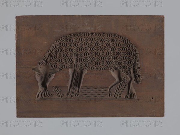 Double-sided gingerbread form; rectangular shape. One side: image of a sheep grazing in a meadow. Other side: figure of a goat with lush fur and long horns curved backwards; at the bottom a fragment of a flowery meadow. Gingerbread; plant ornaments, floral decorations; applied art with zoomorphic motifs and folk motifs. 18th–19th century, Province of Silesia 1742–1918. Height 19.2 cm, width 13.2 cm, depth 3.7 cm., A carved wooden panel with a dark brown surface features a bas-relief of an animal facing left with a textured curly coat, slender legs, and a lowered head near decorative plant shapes carved into the base; the background around the relief is a smooth brown expanse, and the image also shows a pale gray border surrounding the panel.