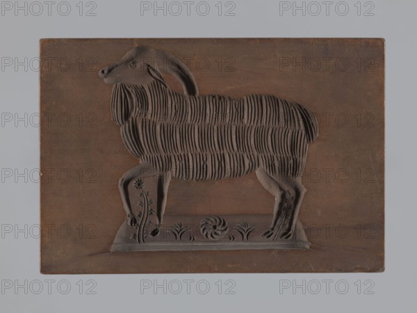 Double-sided gingerbread form; rectangular shape. One side: image of a grazing sheep in a meadow. Other side: goat with lush fur and long backward-curving horns, with a fragment of a flowery meadow at the bottom. Gingerbread mold, applied art with plant ornaments, floral decorations and zoomorphic motifs. 18th–19th century, Silesia Province of Silesia, 1742–1918. Height 19.2 cm; width 13.2 cm; depth 3.7 cm., A carved wooden plaque depicting the silhouette of a sheep standing on a rectangular base, with carved fur texture shown as vertical grooves across its body and decorative floral motifs on the base; the plaque and carving are shades of brown set against a light gray background, with subtle darker brown shadows and slight worn marks on the wood.