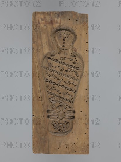 Double-sided gingerbread form. Obverse: female figure in a hat and a long patterned dress. Reverse: baby in swaddling clothes, richly decorated with geometric and plant ornament. Rectangular form; decorative art/folk customs motif. 18th–19th century. Province of Silesia 1742–1918. Height 23.5 cm; width 9.5 cm; depth 2.6 cm., A vertical rectangular wooden plank in brown tones with a carved recessed figure occupying the center, the carving depicting a stylized upright humanlike form with a rounded head, simple facial features, and a hood or hair outline, the torso area filled with repeating leaflike and geometric carved patterns and a bowlike carved element near the lower portion above a floral motif, the wood showing darker and lighter brown grain variations, small round holes scattered across the surface, a few surface cracks and worn edges, and a neutral gray background.
