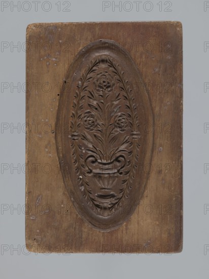 Double-sided gingerbread mould. Obverse: vase with flowers in an oval frame surrounded by a wreath of leaves. Reverse: rectangular recess with a plant ornament, rose motif and initials E and G. Rectangular shape; mould. 18th–19th century, Province of Silesia 1742–1918. Height 16.5 cm, width 11.5 cm, depth 2.7 cm., A rectangular wooden panel with a carved oval medallion at its center containing a symmetrical floral motif; the carving shows stylized flowers and leaves rising from a decorative vase or urn, with flowing curved elements and a textured carved border around the oval. The wood shows a range of brown tones with lighter and darker areas, small pale spots, and worn edges, and the background behind the panel is a muted gray.