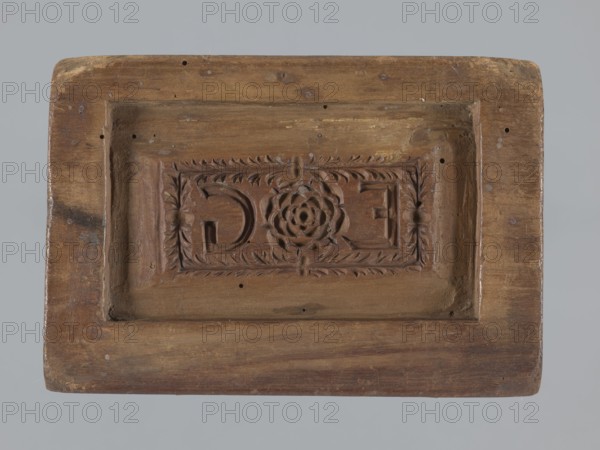 Double-sided gingerbread mould. Obverse: oval frame containing a decorative vase with flowers surrounded by a wreath of leaves. Reverse: rectangular recess with a plant ornament, featuring a rose motif and the initials E and G. Rectangular shape; applied art, folk motifs, floral decorations. 18th–19th century, Province of Silesia 1742–1918. Mould; height 16.5 cm, width 11.5 cm, depth 2.7 cm., A rectangular wooden block with a recessed central panel carved with a floral medallion surrounded by a leafy border, showing worn and roughened surfaces and small holes; the wood displays brown tones with lighter and darker brown variations and some grayish areas, against a pale gray background.