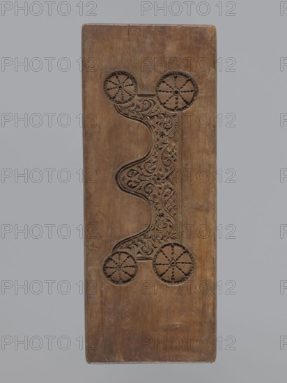 Double-sided gingerbread mould. Side A: figure of a soldier. Side B: various floral and geometric ornaments. Unknown author woodcarver or gingerbread maker. Folk-style decorations; woodcarving; gingerbread mould; applied and decorative art; folk motifs. Double-sided rectangular mould: one side shows a male figure in a Napoleonic-era military uniform, wearing high headgear with a feather, numerous chest decorations, and a plant ornament at the bottom. The other side displays a rich floral and plant ornament enclosed in a tripartite frame, with decorative spoked wheels in the four corners. One edge bears an engraved date 1810 with the letter S between the digits against the background of an anchor the maker's mark. Date: 1810. Place/region: Province of Silesia 1742–1918. Form. Dimensions: height 31.7 cm; width 12.5 cm; depth 2.4 cm., A rectangular wooden panel with a carved central design showing a curving, symmetrical ornamental motif flanked by circular wheel-like carvings at each end;...