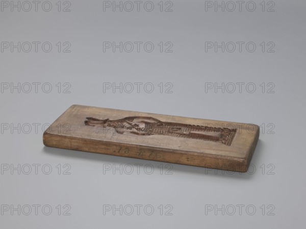 Double-sided gingerbread mould. Side A: figure of a soldier. Side B: various floral and geometric ornaments. Maker unknown woodcarver or gingerbread maker. Folk-style decorations; woodcarving; applied/decorative art; gingerbread moulds; male figures; military uniforms; folk motifs. Description: rectangular double-sided mould — one side shows a male figure in a Napoleonic-era military uniform, wearing a high headgear with a feather and numerous chest decorations, with a plant ornament at the bottom; the other side features a rich floral and plant ornament enclosed in a tripartite frame, with decorative spoked wheels in the four corners. One edge bears an engraved date 1810 with the letter S between the digits against the background of an anchor the maker's mark. Date: 1810. Place: Province of Silesia 1742–1918. Object type: form. Dimensions: height 31.7 cm; width 12.5 cm; depth 2.4 cm., A rectangular wooden block rests on a smooth light gray surface against a pale gray background, showi...