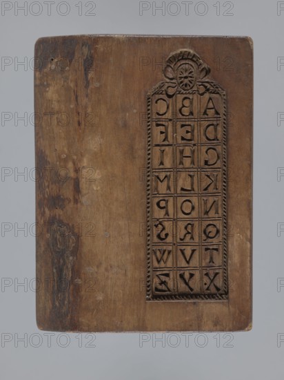 Double-sided gingerbread mould. Obverse: board with letters of the alphabet; reverse: image of a running wild boar. Folk-style decorations; iconography includes wild boar and alphabet. Double-sided rectangular mould; on one side a decorative board with the letters of the alphabet carved on it; the other side: image of a wild boar running across a meadow. On one edge there is a date engraved: 1806, between the digits of the date the letter S on the background of an anchor the mark of the woodcarver or gingerbread maker — the owner of the form. Materials/function: form for gingerbread woodcarving. Date: 1806. Origin: Silesia Province of Silesia, 1742–1918. Dimensions: height 16 cm, width 12 cm, depth 3 cm., A rectangular wooden board with a carved vertical panel near one edge showing a grid of letter-shaped indentations and a decorative floral motif at the top; the wood shows areas of wear and darker stains, with visible grain and small circular marks, and colors include brown, dark brow...