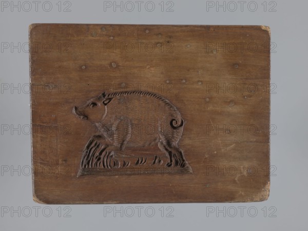 Double-sided gingerbread mould. Obverse: board with letters of the alphabet; reverse: image of a running wild boar across a meadow. Double-sided rectangular mould; on one side a decorative board with the letters of the alphabet carved on it; the other side: image of a wild boar running across a meadow. On one of the edges there is a date engraved: 1806, with the letter S on the background of an anchor mark of the woodcarver or gingerbread maker. Materials/subject: animals, gingerbread, gingerbread moulds, folk-style decorations, wild boar iconographic, alphabet, folk motifs. Date: 1806. Region: Province of Silesia 1742–1918. Type: form. Dimensions: height 16 cm, width 12 cm, depth 3 cm. Creator: unknown., A rectangular wooden board with a carved relief of a pig standing on a textured base; the wood shows a range of brown tones with lighter and darker areas and visible grain, knots, and small circular marks; the pig carving includes carved fur texture, a curled tail, and a raised snout,...