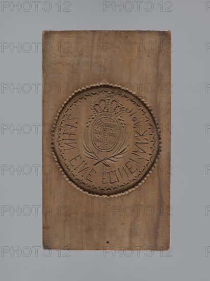 Double-sided gingerbread mould. Obverse: rectangular mould with an oval coat of arms in a decorative round frame topped with a crown; beneath the coat of arms two crossed palm leaves; around it an inscription in German: ZEHN EINE EEINE MARK 1816. Reverse: wreath woven from flowers and ears of grain. Gingerbread mould, folk-style decorative applied art, plant, geometric and floral ornaments. German, Silesia, ca. 1816. Form dimensions: height 15.7 cm, width 9 cm, depth 3.8 cm., A rectangular wooden block with a carved circular emblem in the center, showing a decorative scalloped border and an inner design featuring a crowned oval medallion surrounded by stylized leaves; the wood displays a natural brown color with variations in tone, a few darker brown marks and small spots, and the background around the block is gray.
