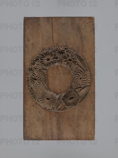 Double-sided gingerbread mould. Rectangular shape; obverse: oval coat of arms in a decorative round frame topped with a crown, two crossed palm leaves beneath, surrounded by a German inscription ZEHN EINE EEINE MARK 1816; reverse: wreath woven from flowers and ears of grain. Gingerbread mould, folk-style applied decorative art, plant, geometric and floral ornaments. Materials/function: mould for gingerbread. Date: ca. 1816. Dimensions: height 15.7 cm, width 9 cm, depth 3.8 cm. Region: Province of Silesia 1742–1918., A rectangular wooden block with a carved circular wreath design near its center; the wood shows a range of brown tones from light to medium with darker brown carved details and grain variations, and the background around the block is a muted gray.