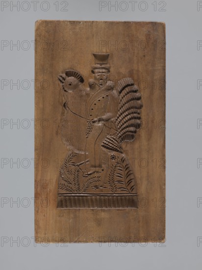 Double-sided gingerbread mold. Obverse: male figure on a rooster; reverse: bird. Double-sided rectangular mold; on one side a male figure in a high hat and fur-lined jacket sitting on a rooster, with a decorative plant-motif border at the bottom; on the other side, a bird facing right with a plant ornament at the bottom. Folk-style decoration; iconography: birds, riders, roosters. Gingerbread mold, 18th–19th century, Province of Silesia 1742–1918. Materials: mold. Dimensions: height 15.5 cm, width 9 cm, depth 3.7 cm., A rectangular wooden panel carved in a brown wood tone shows a relief of a figure standing on stylized foliage, beside and partially overlapping a large bird with a fan-shaped tail; the figure wears a tall hat and clothing with a collar and holds an object near the bird, and the bird has a rounded head and a comb-like crest; the lower area of the carving features carved plants and a horizontal band with vertical grooves; the background around the carved scene is smooth wo...