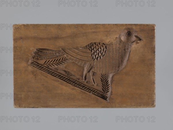 Double-sided gingerbread mold. Obverse: male figure in a high hat and fur-lined jacket sitting on a rooster, with a plant ornament decorative motif at the bottom. Reverse: bird turned to the right with a plant ornament at the bottom. Folk-style figurative scenes; iconography includes birds, riders, and roosters. Mold, rectangular shape, height 15.5 cm, width 9 cm, depth 3.7 cm. 18th–19th century, Province of Silesia 1742–1918., A rectangular wooden panel with a carved bird in profile occupying the center, showing textured feather patterns on its body and layered carved details on its wing and tail, the bird standing on a carved diagonal base that includes leaf-like motifs; the wood has a warm brown tone with variations in shade and a slightly worn surface, set against a neutral gray background.
