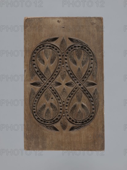 Double-sided gingerbread mould with a plant and floral ornament on both sides;, Gingerbread, woodcarving; folk-style decorations with plant twigs, geometric and plant motifs. Rectangular shape: one side with an elaborate ornament of plaiting and plant and geometric elements; the other side with a decorative motif of two ribbons intertwined in figure-eights filled with and surrounded by lanceolate leaves. 18th–19th century, Province of Silesia 1742–1918. Form. Height 14.5 cm, width 8.5 cm, depth 1.7 cm., A rectangular wooden panel with a carved symmetrical design featuring interlocking looping shapes and leaf-like motifs, the wood surface showing a natural brown color with darker carved recesses and lighter worn areas, against a neutral light gray background.