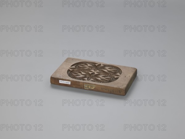 Double-sided gingerbread mould with a plant and floral ornament on both sides;, Gingerbread mould, woodcarving, folk-style decorations; motifs include plant twigs, geometric motifs, plant motifs. Rectangular shape; one side has an elaborate ornament of plaiting with plant and geometric elements; the other side shows two ribbons intertwined in figure-eights filled with and surrounded by lanceolate leaves. 18th–19th century, Province of Silesia 1742–1918. Form. Height 14.5 cm, width 8.5 cm, depth 1.7 cm., A rectangular wooden block with a carved circular floral pattern on its top surface sits on a smooth background; the block is brown with darker brown carved areas and shows natural wood grain and worn edges, and the background is light gray.