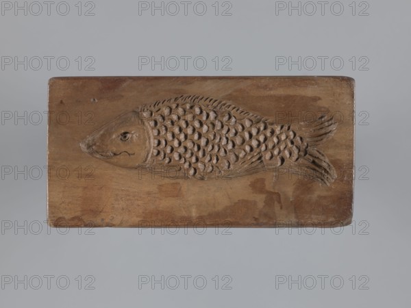 Double-sided gingerbread mould. Obverse: stylised elongated fish covered with scales, shown in profile. Reverse: figure of Archangel Michael in a patterned robe decorated with a plant twig, holding scales in his right hand and a sword in his left, with a plant ornament at the bottom. Christianity; folk-style decorations. Applied decorative art; mould made for gingerbread. Rectangular shape. Silesia Province of Silesia, 1742–1918; 18th–19th century. Materials: unspecified. Dimensions: height 13.5 cm, width 6.5 cm, depth 4.5 cm., A rectangular wooden block with a carved fish in relief occupies the center of the image; the fish carving shows a head with a carved eye and mouth, a textured body of overlapping scale shapes, and a tail with carved fin lines. The wood surface displays a range of brown tones with visible grain and some darker brown patches and worn areas, and the background surrounding the block is a smooth pale gray.