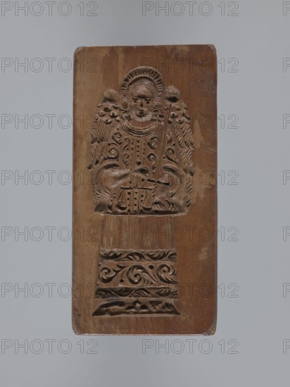 Double-sided gingerbread mould. Rectangular mould; obverse: stylised elongated fish covered with scales, shown in profile; reverse: figure of Archangel Michael in a patterned robe decorated with a plant twig, holding scales in his right hand and a sword in his left, with a plant ornament at the bottom. Christianity, gingerbread, decorative/applied art, folk-style decorations, religious symbols, swords, fish. 18th–19th century, Province of Silesia 1742–1918. Mould. Height 13.5 cm, width 6.5 cm, depth 4.5 cm., A rectangular wooden panel with a carved relief of an angelic figure centered on it, showing a face with a halo, curly hair, detailed wings, and a patterned robe; the figure holds a staff and a pair of scales and stands above a carved decorative base with scrolling motifs. The wood shows natural grain and worn edges, and the visible colors are brown of varying tones and a neutral gray background.