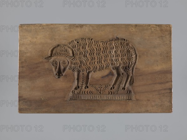 Double-sided gingerbread mold. Obverse: sheep; reverse: hare. Woodcarving, folk-style decorations; decorative and applied art; zoomorphic motifs, gingerbread molds. Rectangular mold: one side shows a sheep standing in a meadow with its head turned forward; the other shows a running hare with a flowery meadow at its feet. Iconography: hares, lamb. 18th–19th century, Province of Silesia 1742–1918. Mold. Height 15.5 cm; width 9 cm; depth 3.6 cm., A rectangular wooden panel with a carved relief of a sheep facing slightly left, showing a textured wavy fleece pattern, curled horns, a stylized face with carved eyes and snout, and slender legs standing on a decorative carved base; the wood surface shows grain variations and small knots, with visible tones of brown and tan on the panel and darker brown in the carved areas, set against a plain gray background.