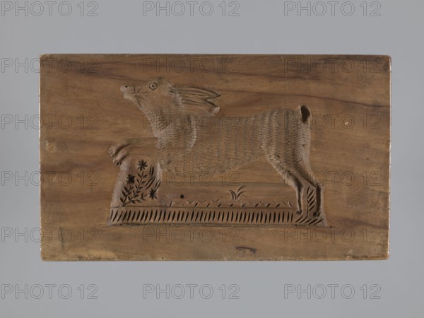 Double-sided gingerbread mold; obverse: sheep, reverse: hare. Woodcarving, folk-style decorations; zoomorphic motifs. Rectangular mold: one side shows a sheep standing in a meadow with its head turned forward; the other shows a running hare above a decorative flowery meadow. 18th–19th century, Province of Silesia 1742–1918. Mold; height 15.5 cm, width 9 cm, depth 3.6 cm., A rectangular wooden panel carved with the raised figure of an animal in profile, appearing to stand on a carved base with decorative floral and linear patterns; the animal has textured fur, a rounded snout, visible carved whiskers, an open circular eye, elongated ears swept back, and a short tail, all rendered in relief against the smooth wood background. The visible colors are various shades of brown in the wood and a neutral gray background surrounding the panel.