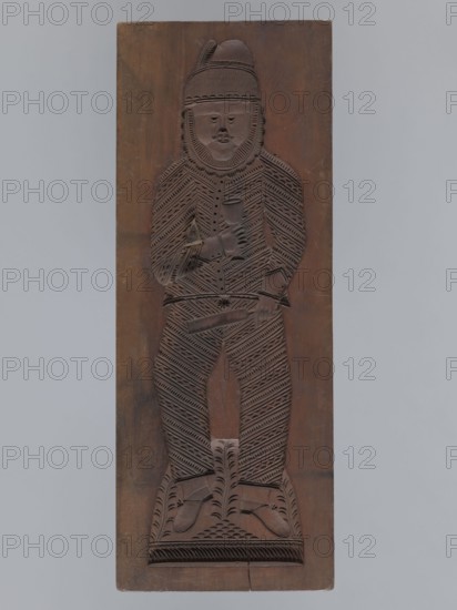 Double-sided gingerbread form. Obverse: male figure with a chalice in hand; reverse: baby in swaddling clothes. Rectangular shape; on one side an image of a male figure with a mustache and beard, wearing a patterned outfit of jacket and trousers, a high hat with a feather, holding a chalice in one hand and a baton, in the other, with a plant ornament at the figure's feet. The other side shows a baby in swaddling clothes, richly decorated with geometric and plant ornaments. Gingerbread form, applied/decorative folk art; folk motifs, plant ornaments. 18th–19th century, Silesia Province 1742–1918. Height 50 cm, width 19 cm, depth 4.5 cm., A tall rectangular wooden panel carved in a brown tone shows a full-length relief of a person wearing a hat with a feather, a patterned long-sleeved garment and patterned leggings; the figure holds a cup in one hand and a flat object in the other, and stands on a carved base with leaf-like motifs; the carving features repeating diagonal and chevron patte...