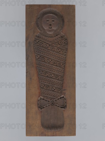 Double-sided gingerbread form. Obverse: male figure with a chalice in hand. Reverse: baby in swaddling clothes. Rectangular shape; on one side an image of a male figure with a mustache and beard, in a patterned outfit consisting of a jacket and trousers, wearing a high hat with a feather, holding a chalice in one hand and a baton, in the other, a plant ornament at the figure's feet. The other side shows a baby in swaddling clothes, richly decorated with geometric and plant ornaments. Gingerbread form, applied/decorative folk art; folk motifs, plant ornaments. Silesia, 18th–19th century. Height 50 cm, width 19 cm, depth 4.5 cm., A carved wooden rectangular panel showing a single stylized human-like figure in relief, with a round face framed by a radiating ruff and simple facial features, a wrapped body covered in repeated diagonal decorative bands of swirl and scallop patterns, a bow-shaped element near the lower part, and a rounded textured area at the bottom; the wood tones range from...