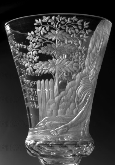 Chalice, glass, c. 1736–1744. Round foot, slightly raised, trace of adhesive; baluster stem, solid, faceted, separated from the foot by two plates with a ring—one plate with a flower—and separated from the bowl by two plates of different diameter and a knob. Conical bowl, twice puffed out at the base, cut into rows of lenses. Engraved decoration covering the entire bowl, biaxial: seated Saint Jerome holding a skull, with a lion at his feet, in a room with bookcases and a side table; inscription: THE LORD'S GIFT OF HIS LIFE / FROM WHICH HE MAKES / THE PRINCE OF SACRIFICES / MAY THE HOUR Strike for so many prosperous years / HOW MANY DROPS IN THE GOBLET / WHAT WINE IS IN THE WINE. Wide geometric-patterned band on the foot. Incised dedication names: Hieronim, Hieronim Florian 1715–1760, Radziwill; religious scenes and dedications. Naliboki, Belarus Grand Duchy of Lithuania context, glassworks production, height 17.2 cm, width 7.7 cm., A clear glass vessel with an etched scene covering its...