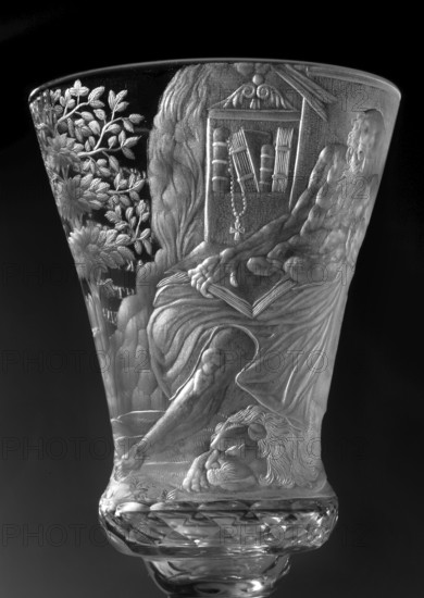 Chalice, glass, 1736–1744. Height 17.2 cm; width 7.7 cm. Round foot, slightly raised, trace of adhesive; baluster stem, solid, faceted, separated from the foot by two plates with a ring one with a flower, separated from the bowl by two plates of different diameter and a knob. Conical bowl, twice puffed out at the base, cut into rows of lenses. Engraved decoration over the entire bowl, biaxial: figure of a seated Saint Jerome holding a skull, a lion at his feet, in a room with bookcases and a side table; inscription in the bowl: THE LORD'S GIFT OF HIS LIFE / FROM WHICH HE MAKES / THE PRINCE OF SACRIFICES / MAY THE HOUR Strike for so many prosperous years / HOW MANY DROPS IN THE GOBLET / WHAT WINE IS IN THE WINE. Foot with a wide strip of geometric pattern. Made in Naliboki, Belarus, Grand Duchy of Lithuania region; dated 1736–1744. Associated names: Hieronim Saint, Hieronim Florian 1715–1760, Radziwill family., A clear decorative glass vessel with detailed raised engravings showing a re...