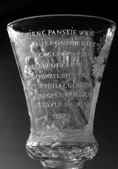 Chalice, glass, 1736–1744. Naliboki, Belarus Grand Duchy of Lithuania. Height 17.2 cm; width 7.7 cm. Round foot, slightly raised, trace of adhesive; baluster stem, solid, faceted, separated from the foot by two plates with a ring, one plate with a flower; separated from the bowl by two plates of different diameter and a knob. Conical bowl, twice puffed out at the base, cut into rows of lenses. Engraved, biaxial decoration covering the entire bowl: seated Saint Jerome holding a skull, a lion at his feet, in a room with bookcases and a side table; inscription in the bowl: THE LORD'S GIFT OF HIS LIFE / FROM WHICH HE MAKES / THE PRINCE OF SACRIFICES / MAY THE HOUR Strike for so many prosperous years / HOW MANY DROPS IN THE GOBLET / WHAT WINE IS IN THE WINE. Religious scenes and dedications; motifs include lions, books, skull anatomist. Associated names: Hieronim Saint; Hieronim Florian 1715–1760; Radziwill family., A close-up view of a clear glass vessel with ornate carved patterns, showin...