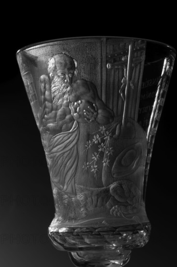 Chalice, Naliboki glassworks 1722–1862. Engraved glass chalice, 1736–1744. Round slightly raised foot with wide strip bearing a geometric pattern; trace of adhesive. Baluster stem, solid, faceted, separated from the foot by two plates with a ring one plate with a flower, and from the bowl by two plates of different diameter and a knob. Conical bowl, twice puffed out at the base, cut into rows of lenses. Engraved, biaxial decoration over the entire bowl: seated Saint Hieronim Saint Jerome holding a skull, with a lion at his feet, in a room with bookcases and a side table; inscription: THE LORD'S GIFT OF HIS LIFE / FROM WHICH HE MAKES / THE PRINCE OF SACRIFICES / MAY THE HOUR Strike for so many prosperous years / HOW MANY DROPS IN THE GOBLET / WHAT WINE IS IN THE WINE. Religious scenes and dedications; references to Hieronim Florian 1715–1760 and the Radziwill family. Origin: Naliboki, Belarus Grand Duchy of Lithuania context. Vessel. Height 17.2 cm; width 7.7 cm., A close-up of a carved...