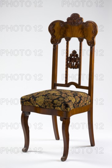 Mahogany chair with a padded seat and a high openwork backrest, with carved decoration. Square legs, S-shaped at the front with volute feet, and arched rear legs. Wavy frame with rounded front corners, with a seat in the form of an overlay cushion. Backrest framed on the sides by straight stiles extending the rear legs, topped with a board with a symmetrical, wavy-cut outline, accentuated by carved acanthus appliqués. The filling has two straight stiles covered with astragalus, topped and fastened together with acanthus leaf arrangements, and a narrow lower stile, arched between the stiles. Plant decoration: acanthus ornament; Louis Philippe style. Dated 1850–1860, Poland Warsaw?. Seating furniture; height 118.7 cm, width 51 cm, depth 53.7 cm., A wooden chair with a carved top rail and slender vertical supports, showing a medium to dark brown polished finish, featuring carved floral motifs on the backrest and a decorative turned central splat, with a padded seat upholstered in a patter...